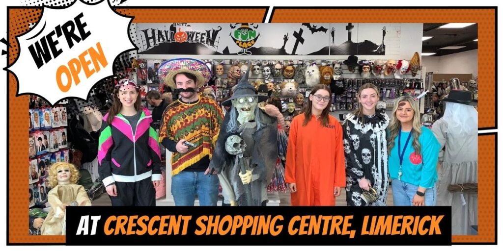 A group of five staff members dressed in colourful Halloween costumes, standing inside a Halloween-themed store with decorations, masks, and props on display. A large banner says ‘We’re Open’ and text below reads ‘At Crescent Shopping Centre, Limerick.