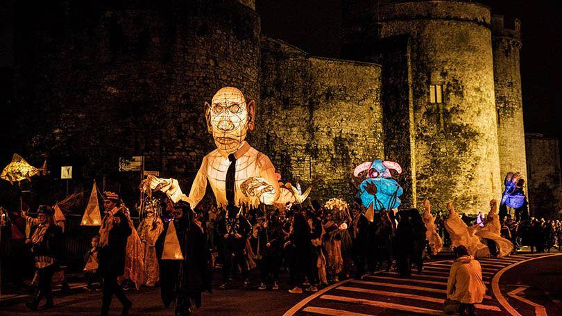 A vibrant nighttime parade at Lumen Street Theatre’s ‘Samhain: Limerick’s Hallowe’en Festival,’ featuring illuminated giant puppets and costumed performers in front of a historic stone castle. Photo by Brian Arthur